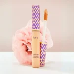 A tube of Tarte shape tape full-coverage concealer - 12B fair beige stands against a soft pink flower. Its cap and base have a vibrant purple and gold chevron pattern.