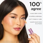 A woman with long hair applies makeup with a brush, showcasing Tarte Shape Tape Contour Concealer in Light Sand.