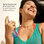 A smiling woman holds a bottle of Sol de Janeiro Cheirosa Perfume Mist from the Tropical Mini Fragrance Collection.